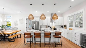A modern kitchen with white cabinets, a central island with bar stools, pendant lighting, and a dining area with a wooden table and chairs. - Hogan Crest w/Heated Pool & Optional Boat Rental (Mooresville)