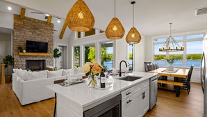 A modern kitchen with a large island, white cabinetry, and a stone fireplace. There is a dining area with a wooden table and chairs, and a view of the outdoors through large windows. - Hogan Crest w/Heated Pool & Optional Boat Rental (Mooresville)