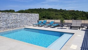 A rectangular swimming pool with clear blue water, surrounded by light-colored tiled flooring. There are several blue lounge chairs arranged around the pool, and a stone wall with a grid pattern on one side. - Porto Vecchio villa with private heated pool near Saint Cyprien beach (Porto-Vecchio)