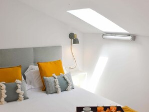 Un dormitorio moderno con tragaluz, una cama con múltiples almohadas, una mesita de noche con lámpara y una bandeja con alimentos para el desayuno.