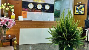 Zona de recepción de hotel con mostrador de madera, un reloj que muestra Nueva York, Hanói y Seúl, y una planta verde grande en primer plano.