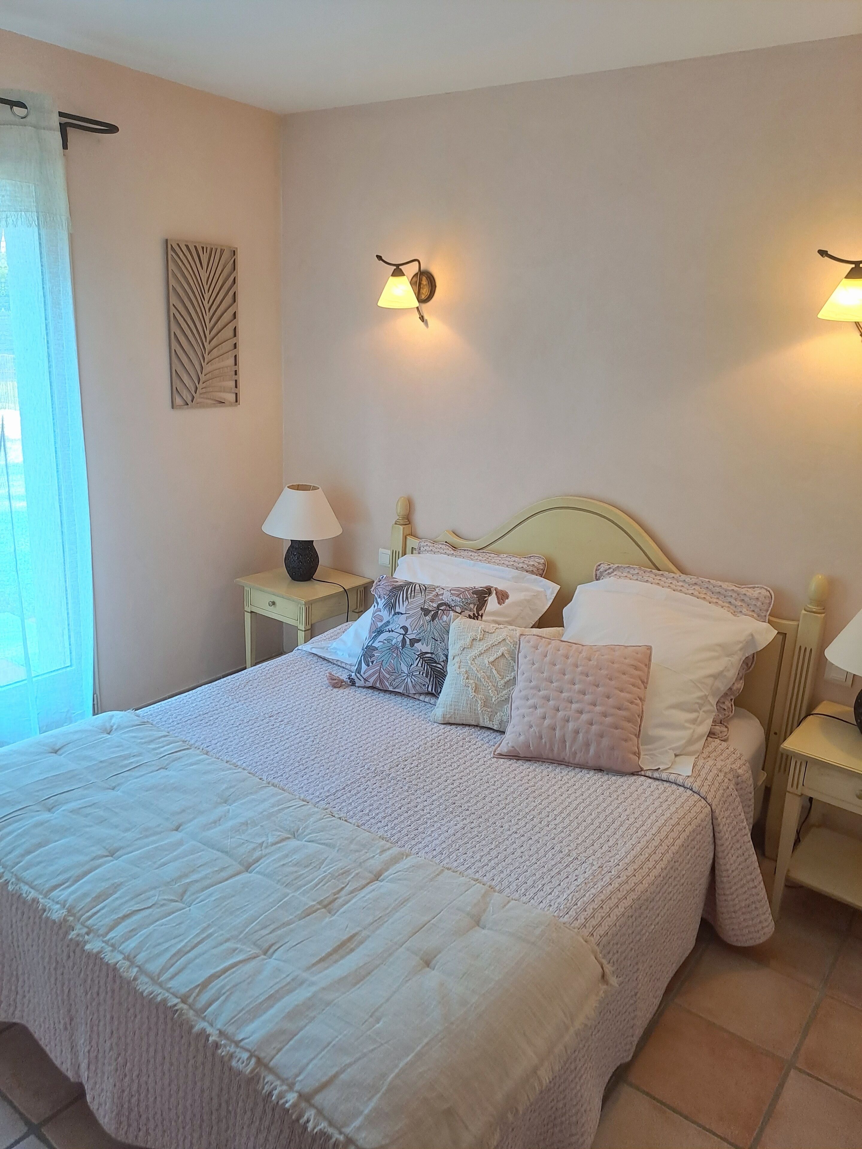 Een net opgemaakt bed met een sprei en meerdere kussens, twee nachtlampjes, een decoratief object aan de muur, en een raam met gordijn.