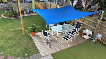 A backyard deck with a table and chairs, surrounded by a wooden fence and a blue shade sail.