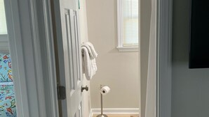 Een badkamer met een witte deur, een handdoekrek met handdoeken, een vloerlamp, een raam met zonwering en een blauw vloerkleed.