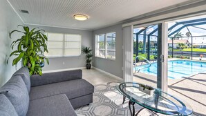 Ein modernes Wohnzimmer mit einem grauen Ecksofa, einem Glastisch und Blick auf einen Pool im Freien.