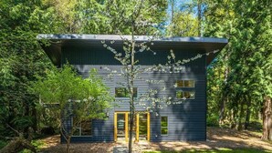 Una casa moderna con facciata scura, circondata da alberi fitti e un piccolo giardino con una panchina.