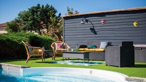 Un espace piscine doté d’une terrasse en bois sombre, d’un canapé en osier et d’un fauteuil en osier.