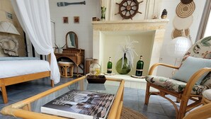 Un salon chaleureux avec un lit, un fauteuil en osier, une table basse avec un livre et une cheminée agrémentée d’objets décoratifs.