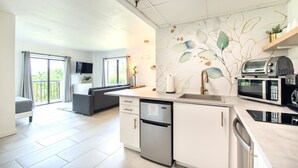 A modern kitchen with a dishwasher, microwave, and toaster. There is a dining area with a sofa and a TV. The room has a balcony with a view of greenery. - Lovely Studio near Universal Studios (Orlando)