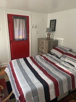 Une chambre avec une porte rouge, une commode en bois, un lit avec une literie à rayures et une lampe sur la commode.