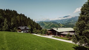 Un paysage rural avec une maison et une grange, entourés d’une végétation luxuriante et avec des montagnes en arrière-plan.