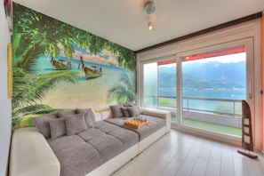 Un salon moderne agrémenté d’une grande fresque représentant une plage tropicale, d’un canapé gris avec des coussins, d’un jeu d’échecs et d’une grande porte-fenêtre coulissante donnant sur un balcon avec vue sur les montagnes et l’eau.