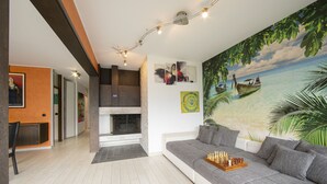 A modern living room with a grey sofa, a fireplace, a chessboard, and a tropical beach mural. - Melide On The Lake for 4, Melide, Switzerland (Melide)