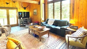 A spacious living room with a high ceiling, wooden walls, a large window, a sofa, a coffee table, and a television.