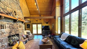 A spacious living room with a stone fireplace, wooden ceiling, and large windows.