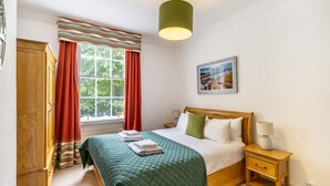 Een slaapkamer met een bed, nachtkastje, lamp, raam met gordijnen, schilderij aan de muur en een kledingkast.