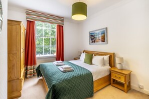 A bedroom with a bed, a nightstand, a lamp, a window with curtains, a painting on the wall, and a wardrobe. - 9 St John's Street -  a family break that sleeps 6 guests  in 3 bedrooms (York)