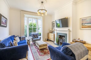 A living room with a blue sofa set, a fireplace, a television, and a large window with a view of greenery. - 9 St John's Street -  a family break that sleeps 6 guests  in 3 bedrooms (York)
