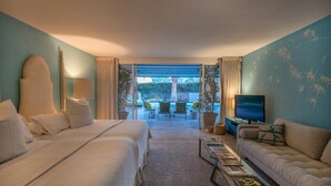 Ein Hotelzimmer mit Bett, Sofa, Fernseher und einem Glastisch. Blick auf einen Außenbereich mit Stühlen und einem Pool.