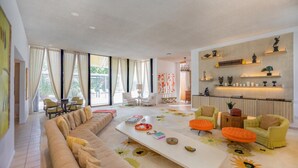 Ein modernes Wohnzimmer mit einer großen Couch, einem Couchtisch und orangefarbenen Hocker. Es gibt mehrere Sessel, einen Kamin und ein großes Fenster mit Vorhängen.