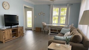 A living room with a TV on a wooden stand, a sofa, a small table with a plant, and a dining area with a table and chairs. - Historic Cottage  (Delray Beach)