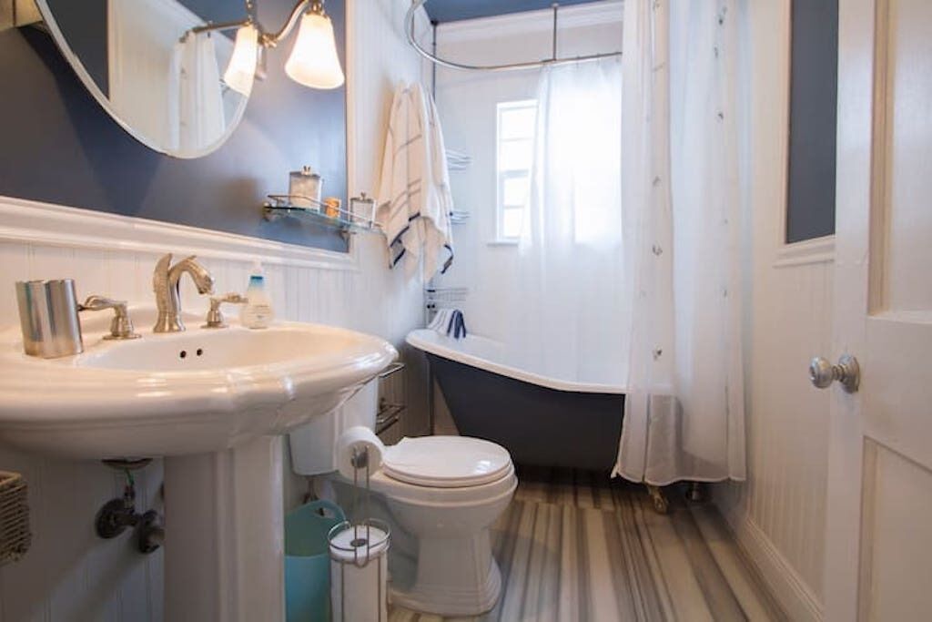 Une salle de bain avec un lavabo, des toilettes et une baignoire, tous de couleur blanche. Un miroir est placé au-dessus du lavabo et il y a une fenêtre avec des rideaux.