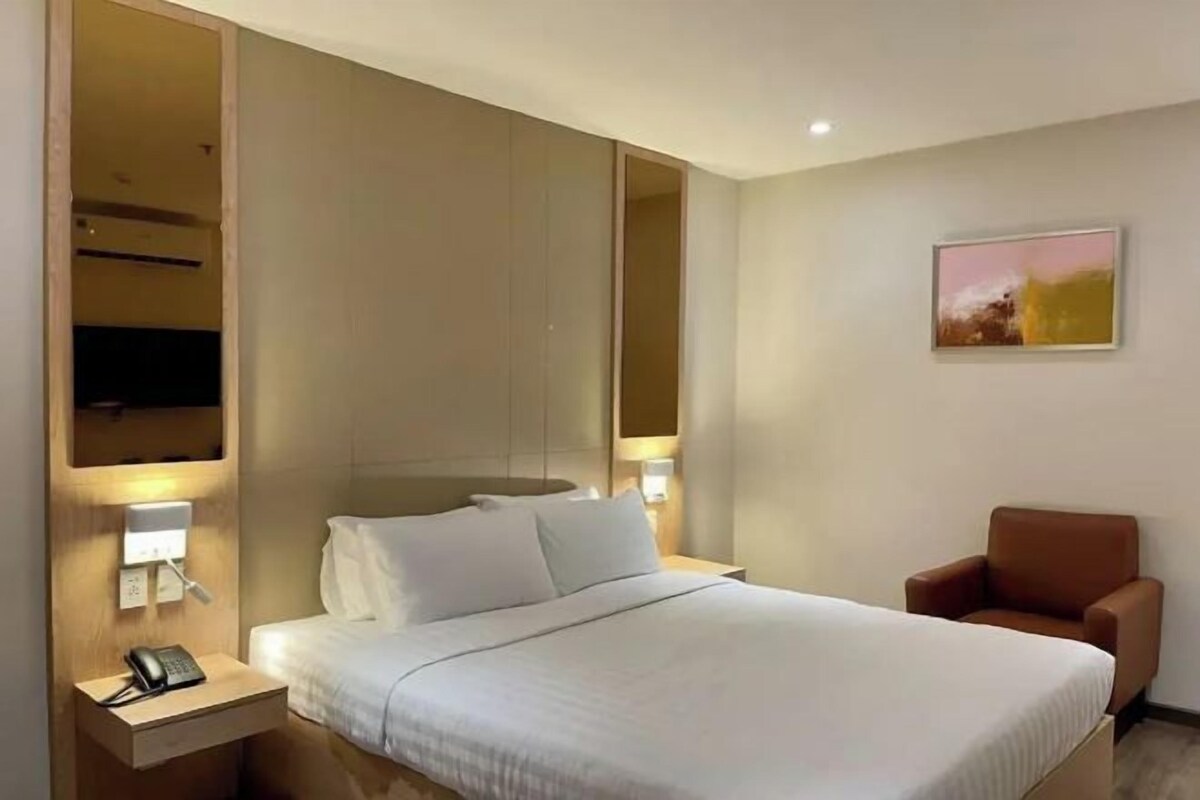 A hotel room with a bed, bedside table with a phone, a chair, a wall-mounted TV, and a painting.