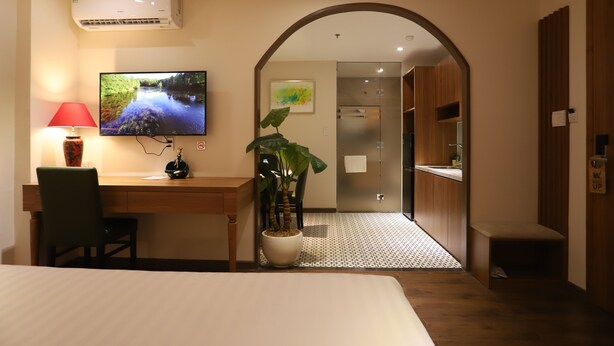 Une chambre d’hôtel avec un lit, un bureau, une télévision, une plante en pot et une vue sur le couloir.