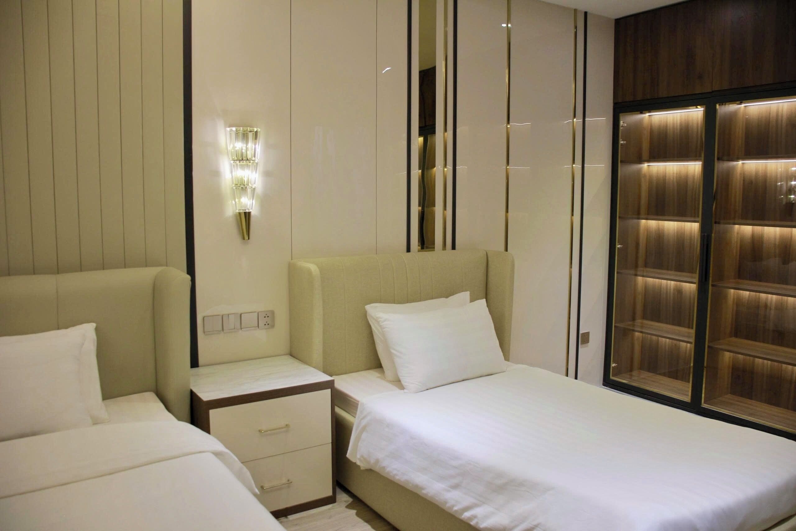 A hotel room with two beds, a nightstand, a wall-mounted light, and a wooden wardrobe.