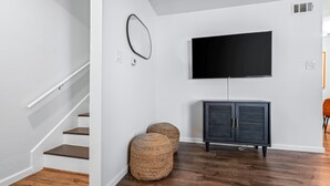 Un salon moderne avec un escalier, un téléviseur à écran plat fixé au mur, un meuble de couleur sombre et deux poufs en fibres tressées.