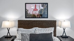Une chambre d’hôtel avec un lit, deux tables de chevet, deux lampes et un tableau représentant une enseigne de motel accroché au mur.