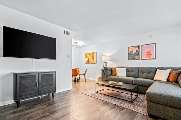 Een moderne woonkamer met een flatscreen televisie, een grijze bank en een eethoek met oranje stoelen.