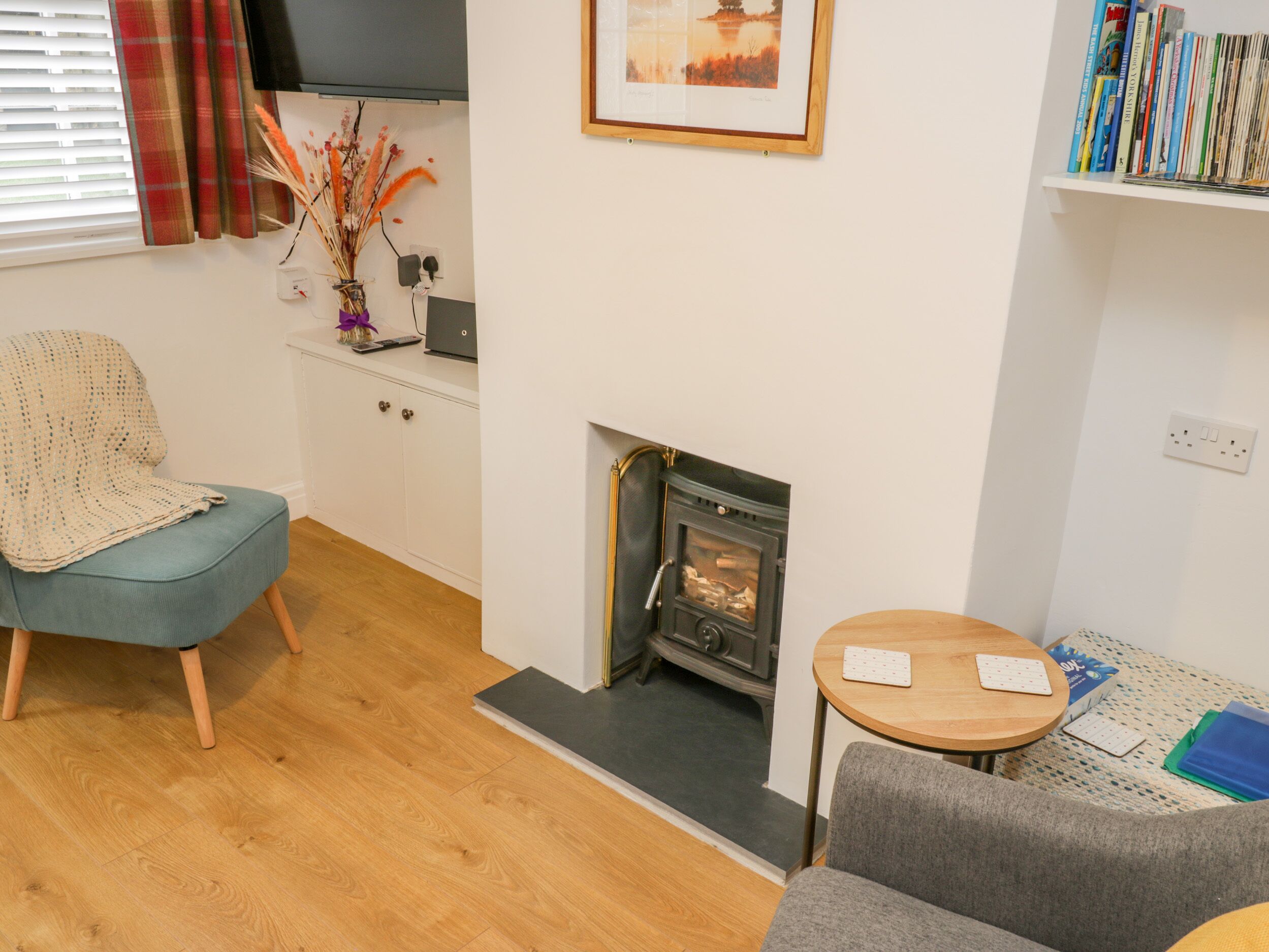 A cozy living room with a fireplace, a blue armchair, a grey sofa, a small wooden table, and a bookshelf with books.
