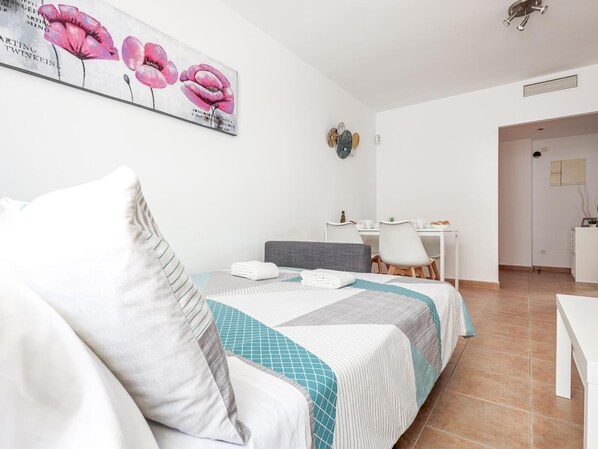 Ein Hotelzimmer mit einem Bett, einem Schreibtisch und einem Stuhl. An der Wand hängt ein Gemälde, und eine Tür führt in einen weiteren Raum.