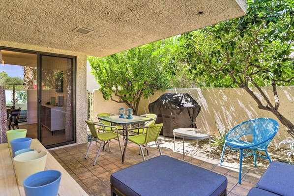 Relax in the serene patio area, complete with a round table, four chairs, and two lounge chairs, surrounded by a large tree and a grill for outdoor cooking.
