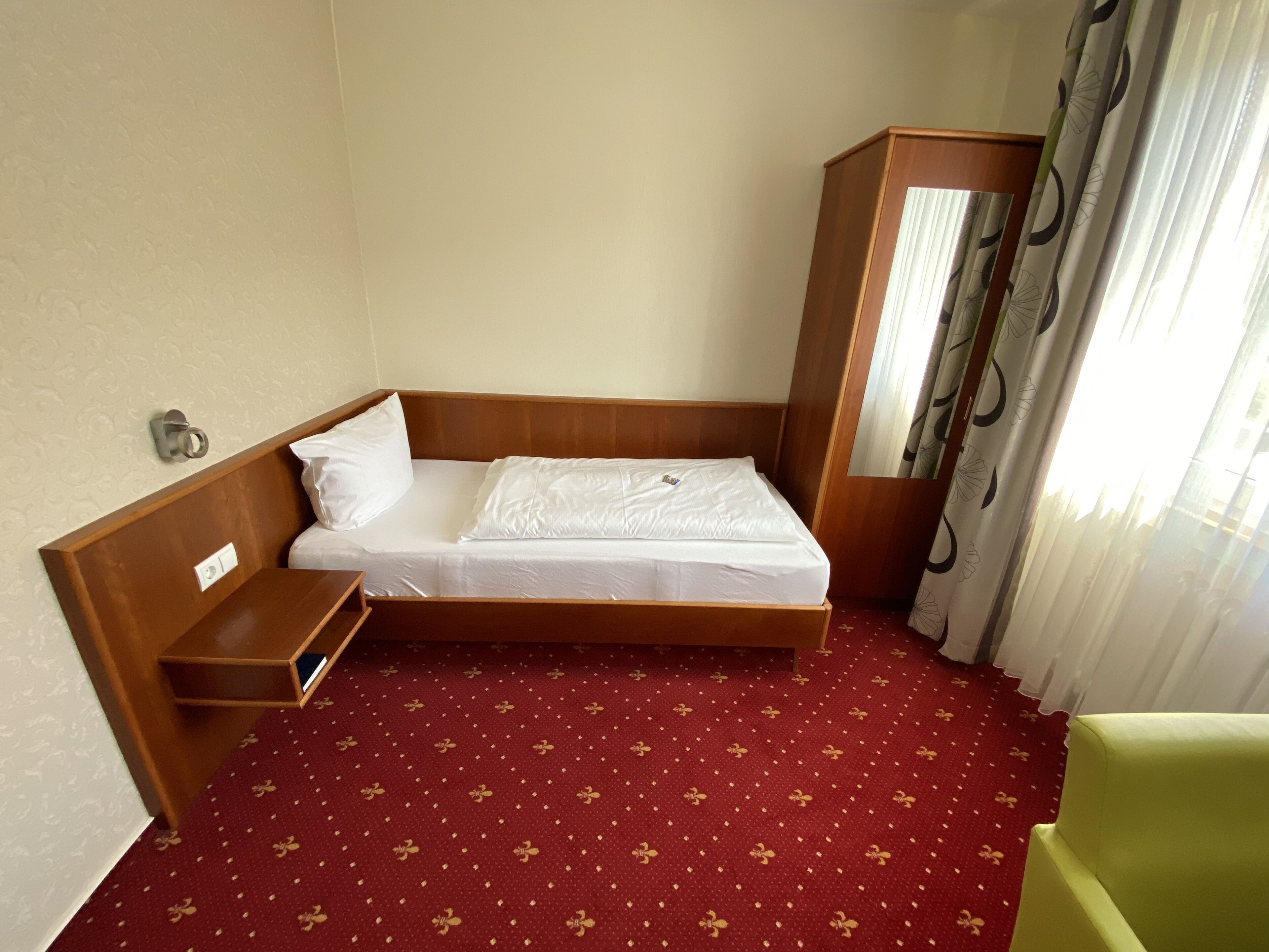 A single bed with a wooden headboard, a bedside table, a red patterned carpet, and a green chair.