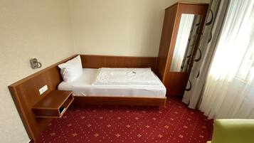 A single bed with a wooden headboard, a bedside table, a red patterned carpet, and a green chair.