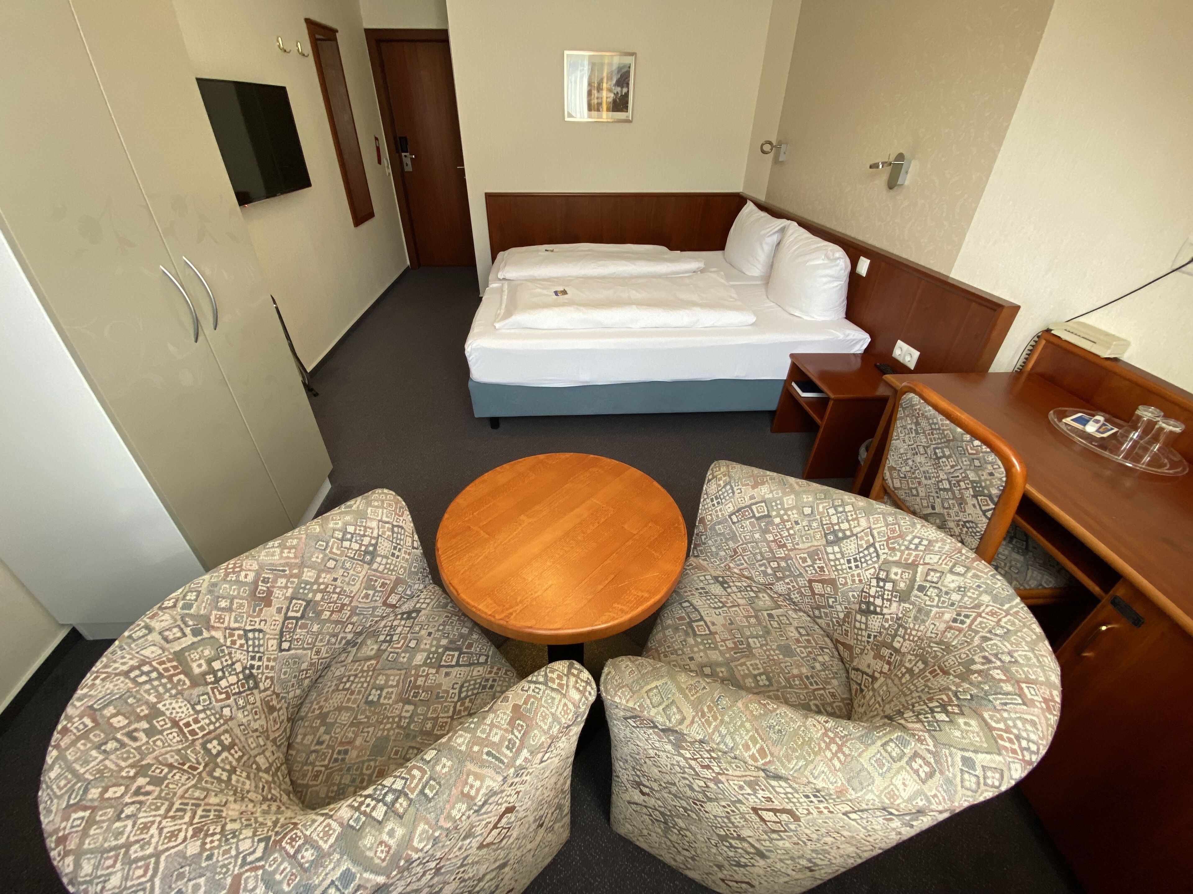 A hotel room with a bed, two patterned armchairs, a small wooden table, a desk with a chair, and a TV mounted on the wall.