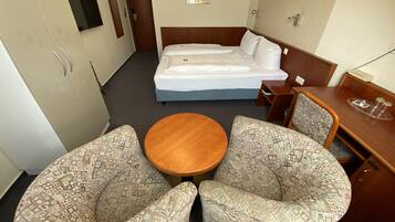 A hotel room with a bed, two patterned armchairs, a small wooden table, a desk with a chair, and a TV mounted on the wall.