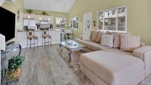 A spacious living room with a beige sofa, a coffee table, and a TV. There is a kitchen area with a dining table and chairs.