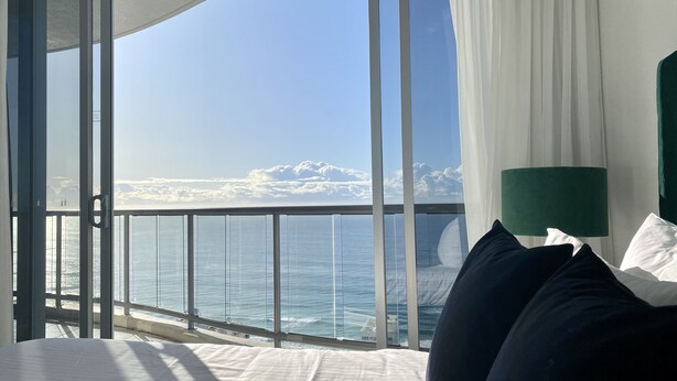 A hotel room with a large window overlooking the sea, a bed with white linens, and a green bedside lamp. - Stunning 3BR, 42nd Floor Skyhome - Breathtaking views!  (Surfers Paradise)