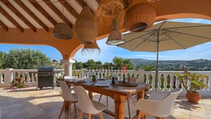 Eine Terrasse im Freien mit einem Holztisch, weißen Stühlen, hängenden Lampen aus Korbgeflecht und einem großen Sonnenschirm.