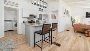 A modern kitchen with white cabinets, a central island with bar stools, a hanging light fixture, and a dining area with a wooden table and chairs. - Remodeled Lakefront! Private beach, toys & hot tub (Little Elm)