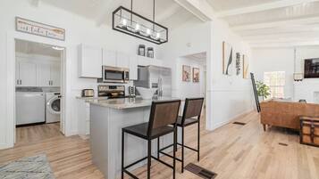 A modern kitchen with white cabinets, a central island with bar stools, a hanging light fixture, and a dining area with a wooden table and chairs.