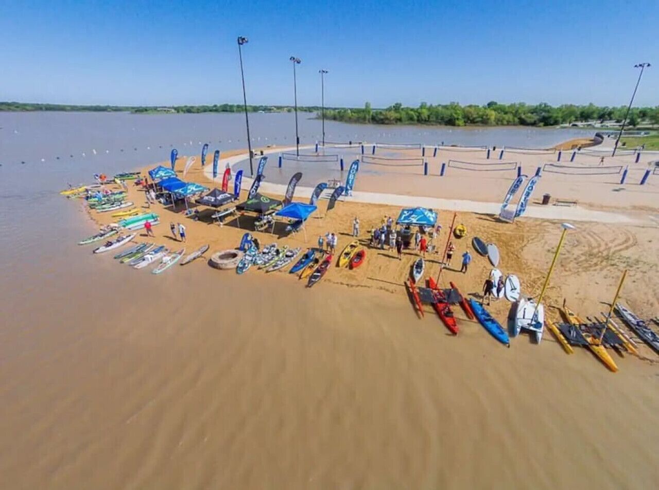 An aerial view of a beach with numerous kayaks and canoes lined up on the sand, a few people standing, and a clear sky.