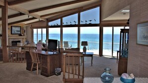 A room with a large window overlooking the ocean, a wooden desk with a computer, a chair, and a small table with a lamp.