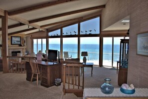 A room with a large window overlooking the ocean, a wooden desk with a computer, a chair, and a small table with a lamp.