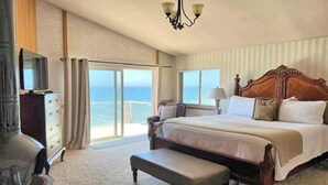 A bedroom with a large bed, a bench, a dresser, a TV, and a view of the ocean.