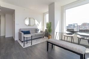 Un salon moderne avec un canapé gris, une table basse ronde, un banc et une grande fenêtre donnant sur la ville.