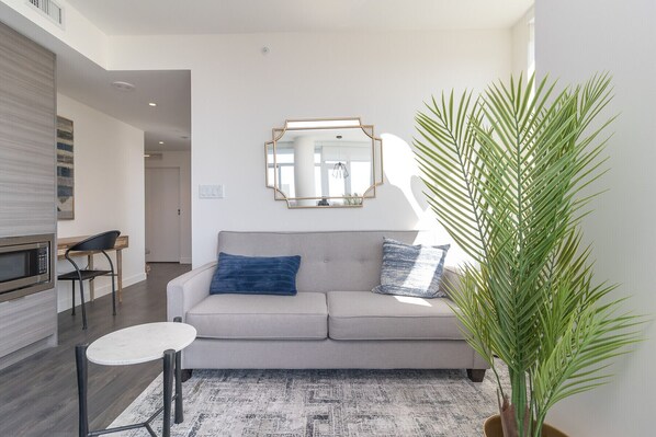 Un salon moderne avec un canapé gris, deux coussins bleus, une petite table blanche et une grande plante en pot.